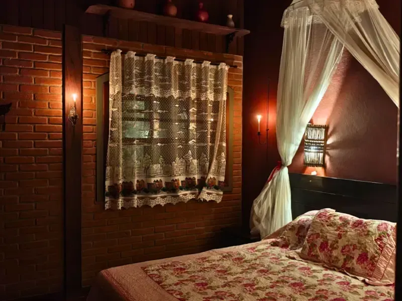 Quarto aconchegante com parede de tijolos vermelhos, cortinas de renda, luz quente de abajur e colcha floral.