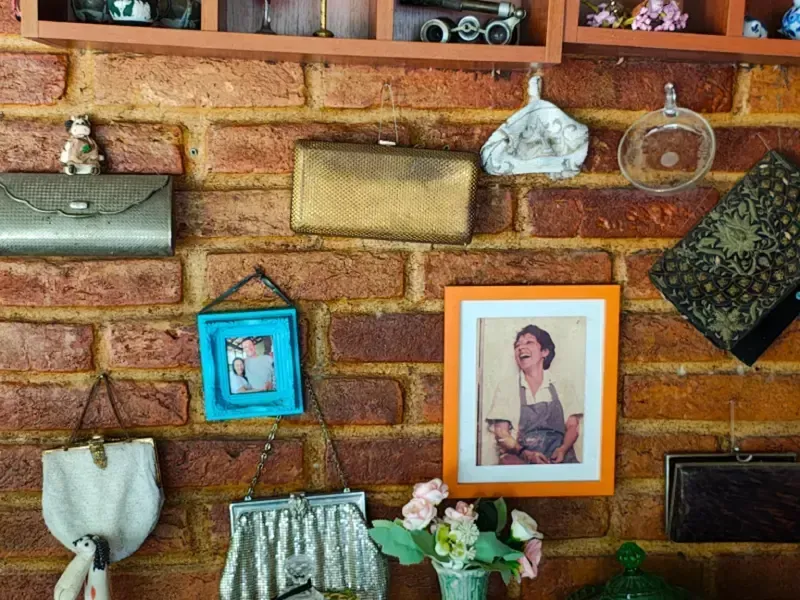 Parede com bolsas penduradas, fotos emolduradas e um pequeno vaso de flores sobre um fundo de tijolos.