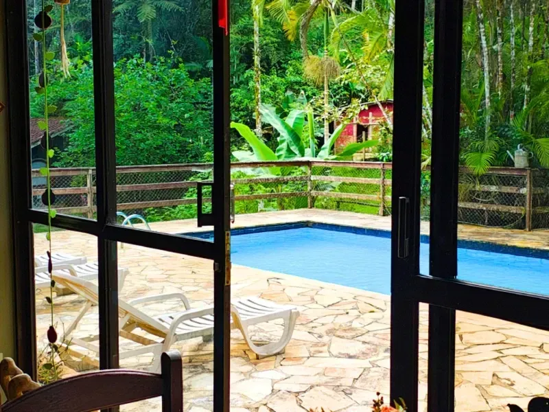 Vista do pátio para uma pequena piscina azul rodeada por espreguiçadeiras e vegetação tropical exuberante.