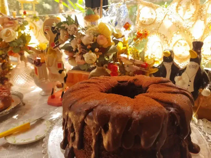 Bolo Bundt de chocolate sobre uma mesa decorada com flores e taças, sob luz quente.