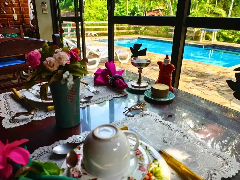 Mesa de café da manhã à beira da piscina, com xícara de chá, flores e doces em um pátio ensolarado.