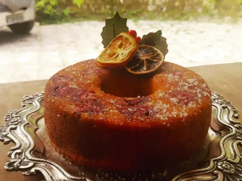 Bolo redondo cor de caramelo servido numa bandeja de prata ornamentada, coberto com fatias de frutas secas.