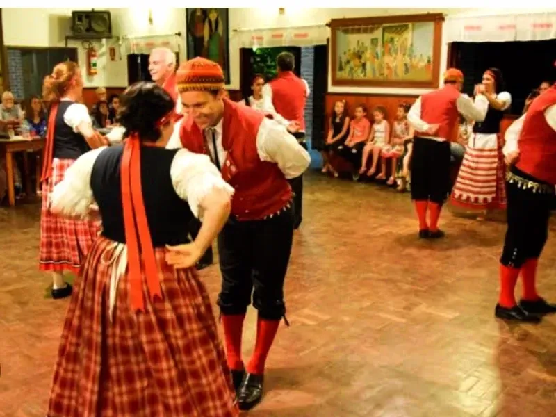 Dançarinos em trajes folclóricos vermelhos e brancos se apresentam em um salão comunitário enquanto o público sentado assiste.