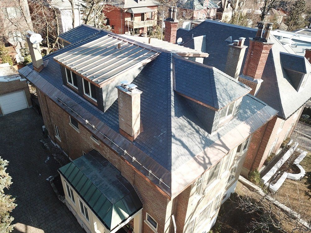 High-angle aerial view of a brick house roof with slate shingles, chimneys, and dormer windows.