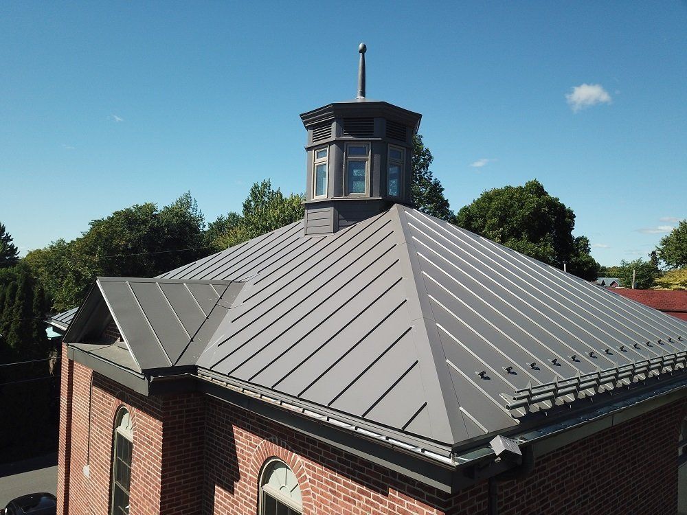 An aerial view of a gray standing-seam metal roof on a brick building with a central cupola and chimney against blue sky.