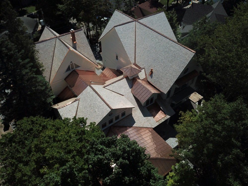An aerial view of a large residential roof with gray shingles and copper metal accents, surrounded by mature trees.
