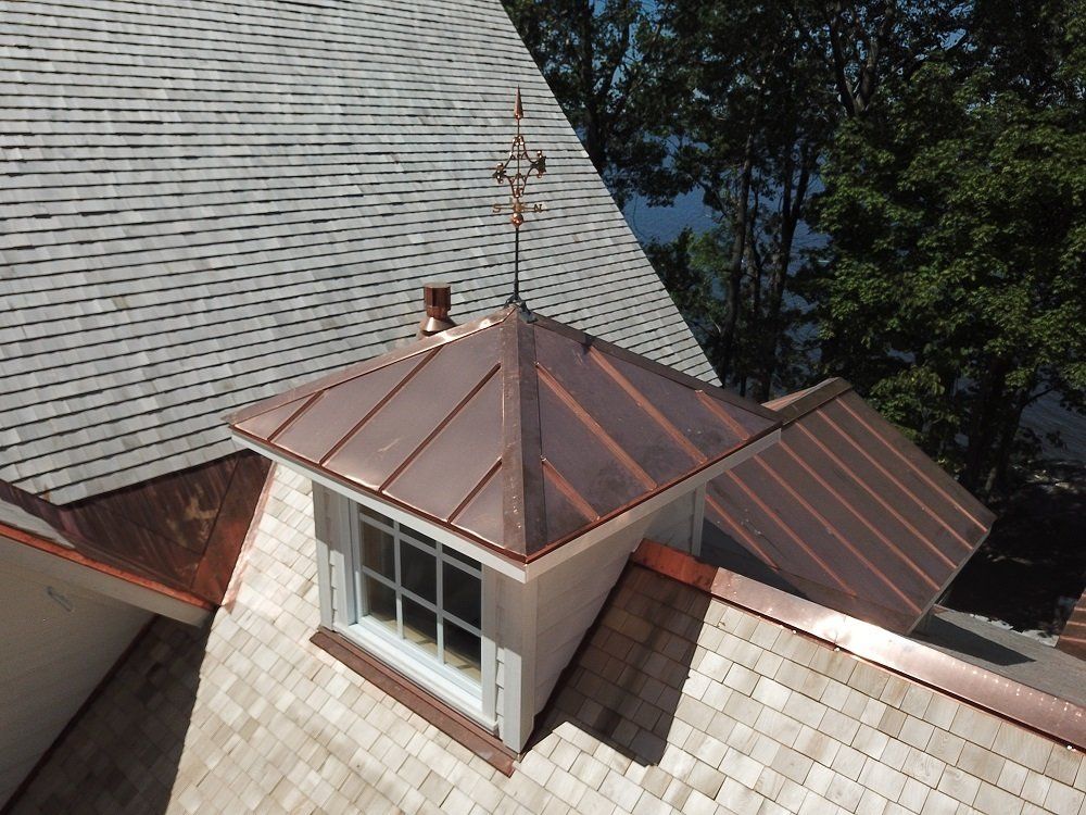 A close-up of a house dormer with a copper roof and finial, set against a gray shingled roof and trees in the background.