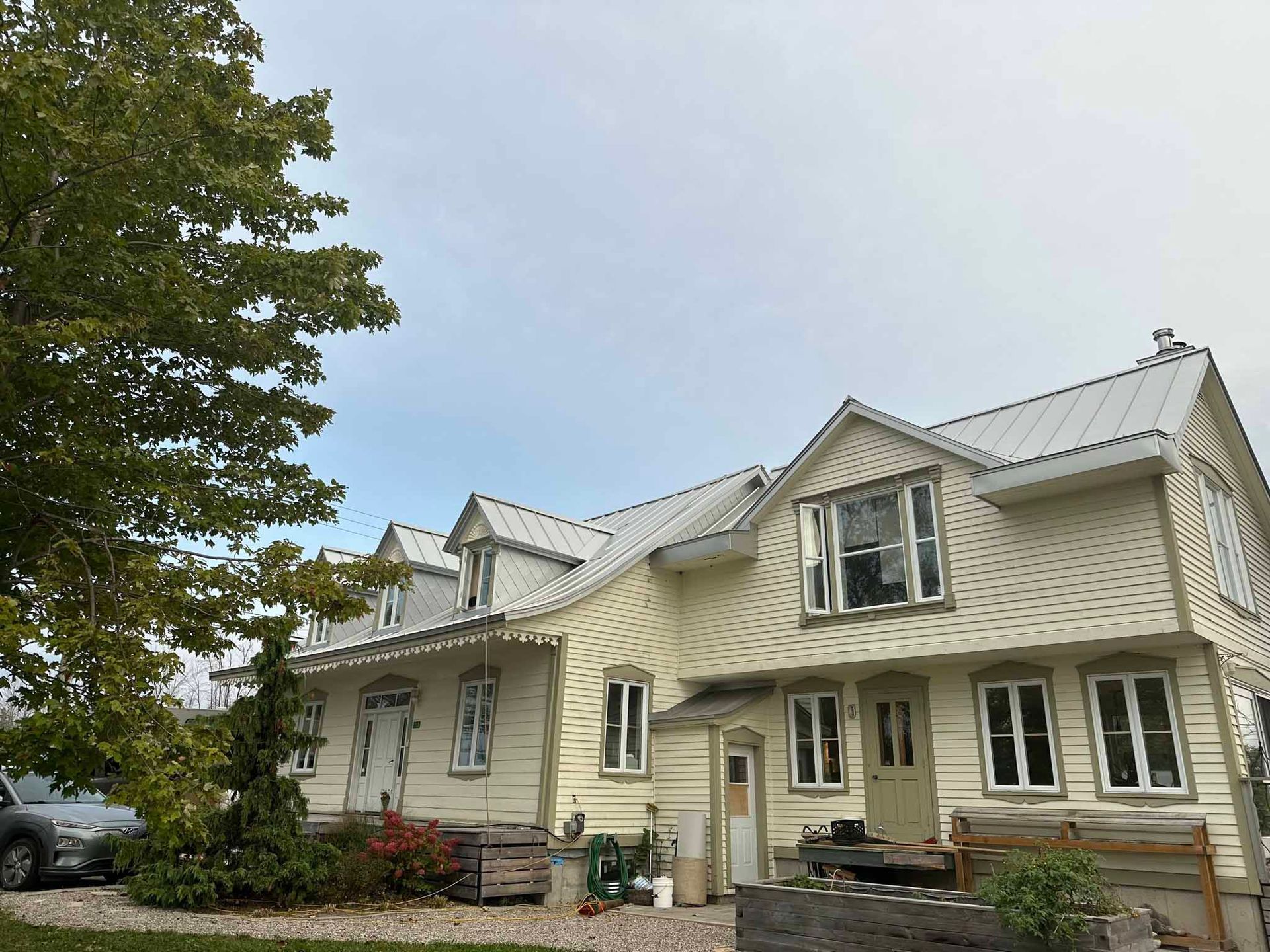 A two-story yellow farmhouse with a white metal roof and dormer windows, featuring a tree on the left and a gravel yard.