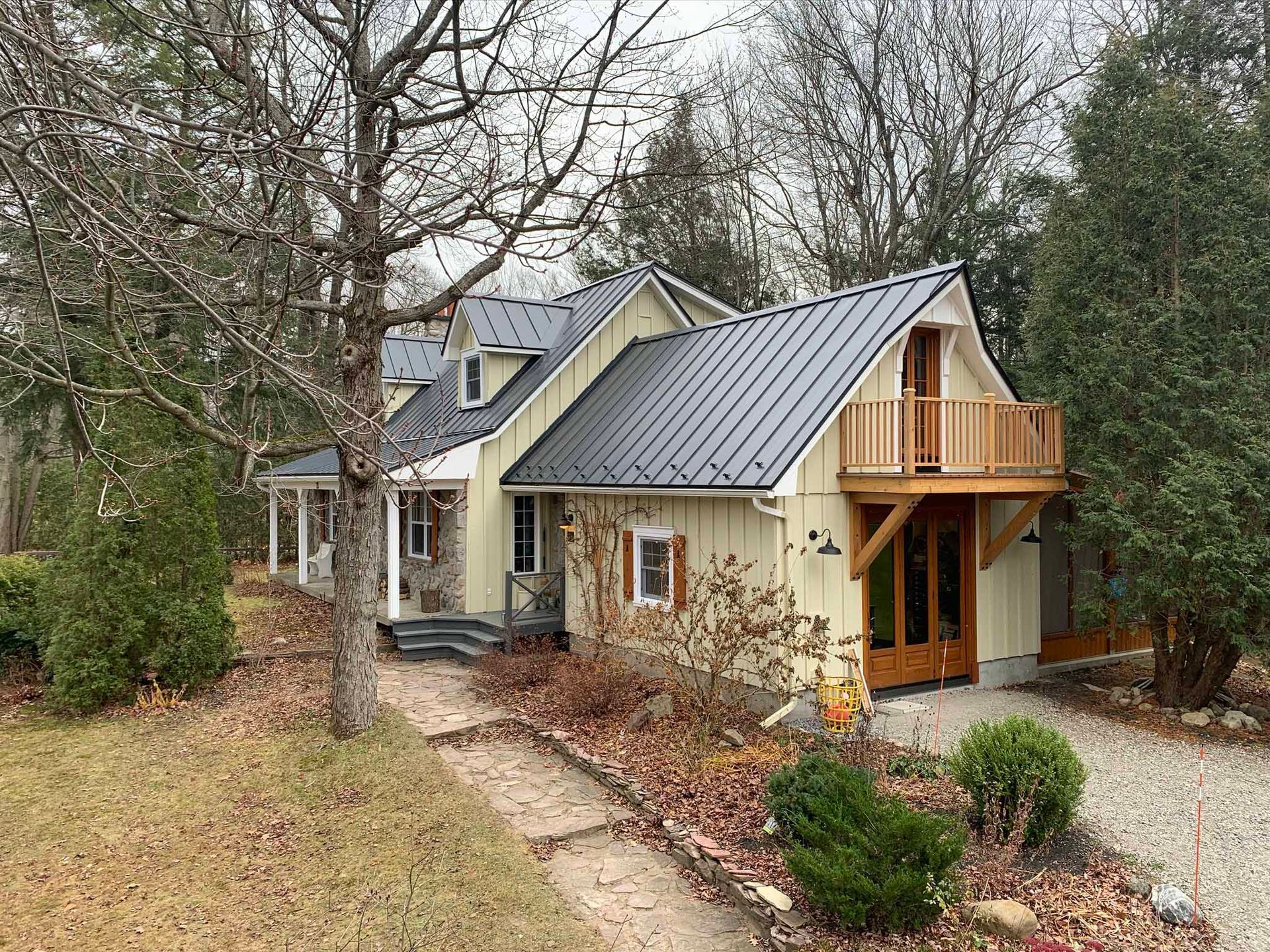 A two-story yellow farmhouse with a dark metal roof, a wooden balcony, and a gravel driveway in a wooded setting.