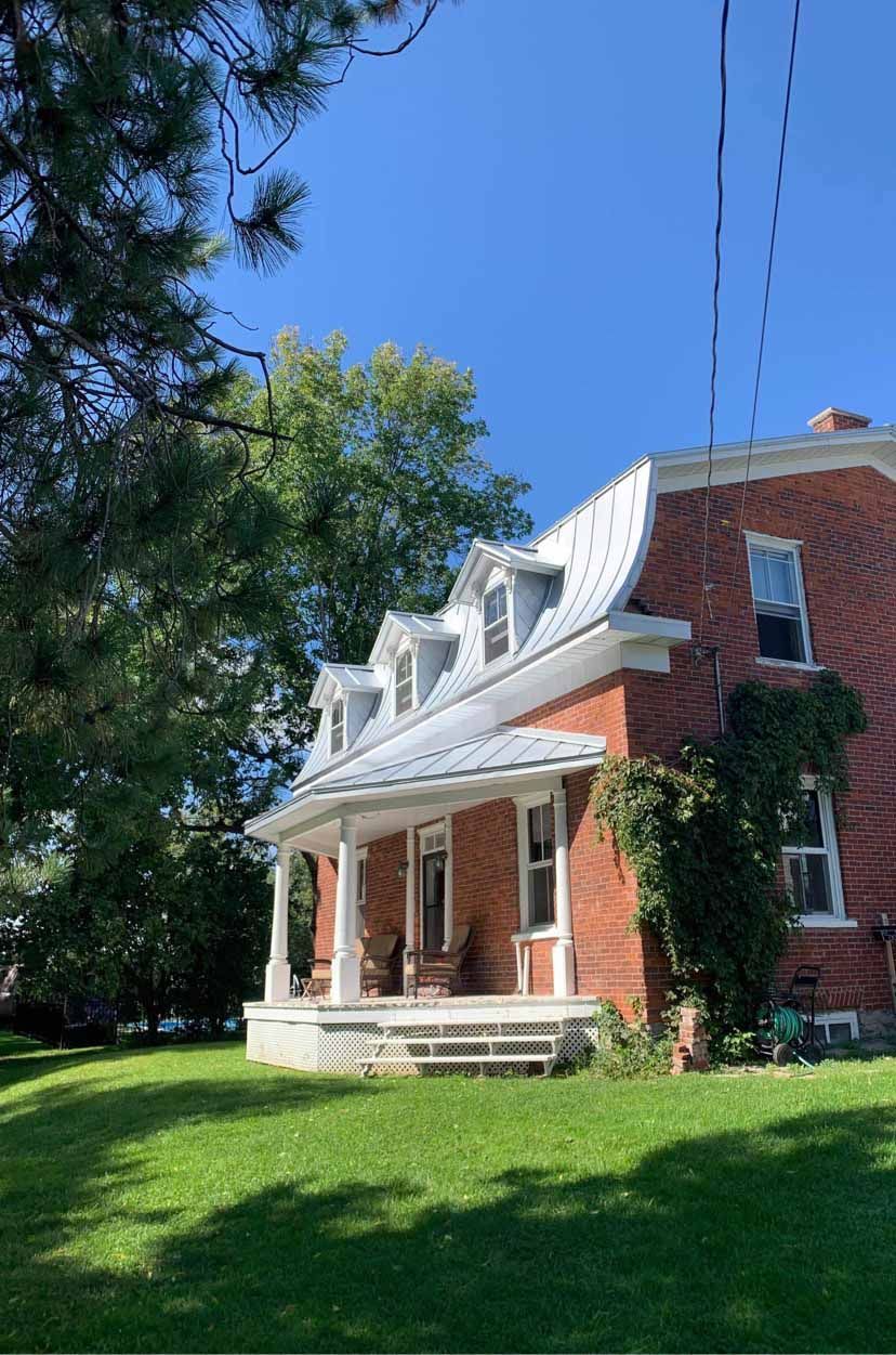 A red brick house with a gambrel roof and front porch sits on a grassy lawn under a clear blue sky.