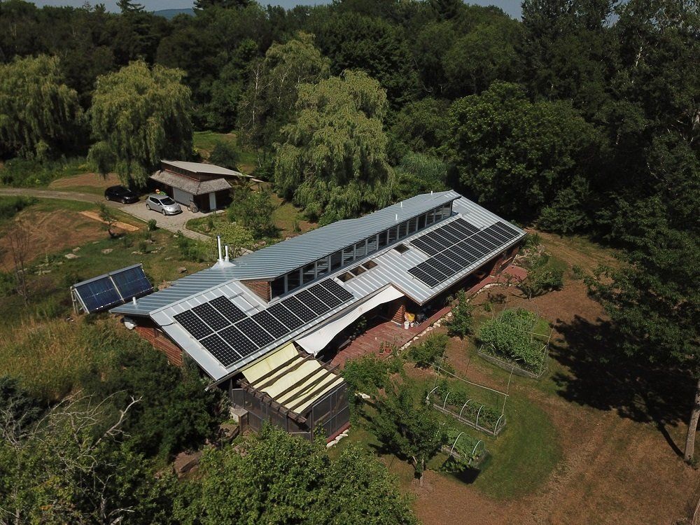 Aerial view of a long, solar-paneled home nestled in a lush, green woodland setting with garden beds.