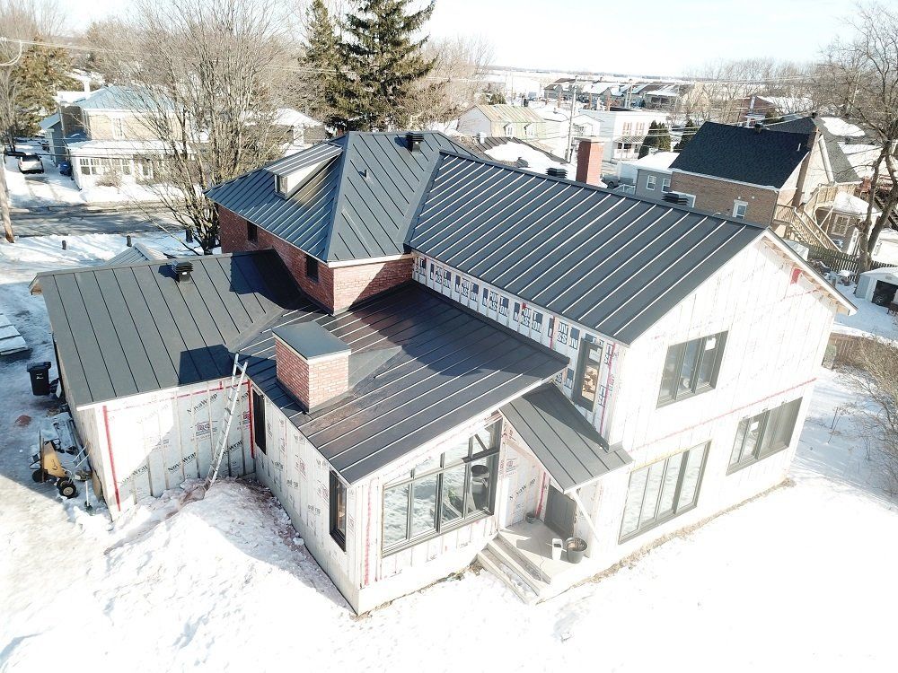 An aerial view of a construction site featuring a house with a newly installed black metal roof in a snowy environment.