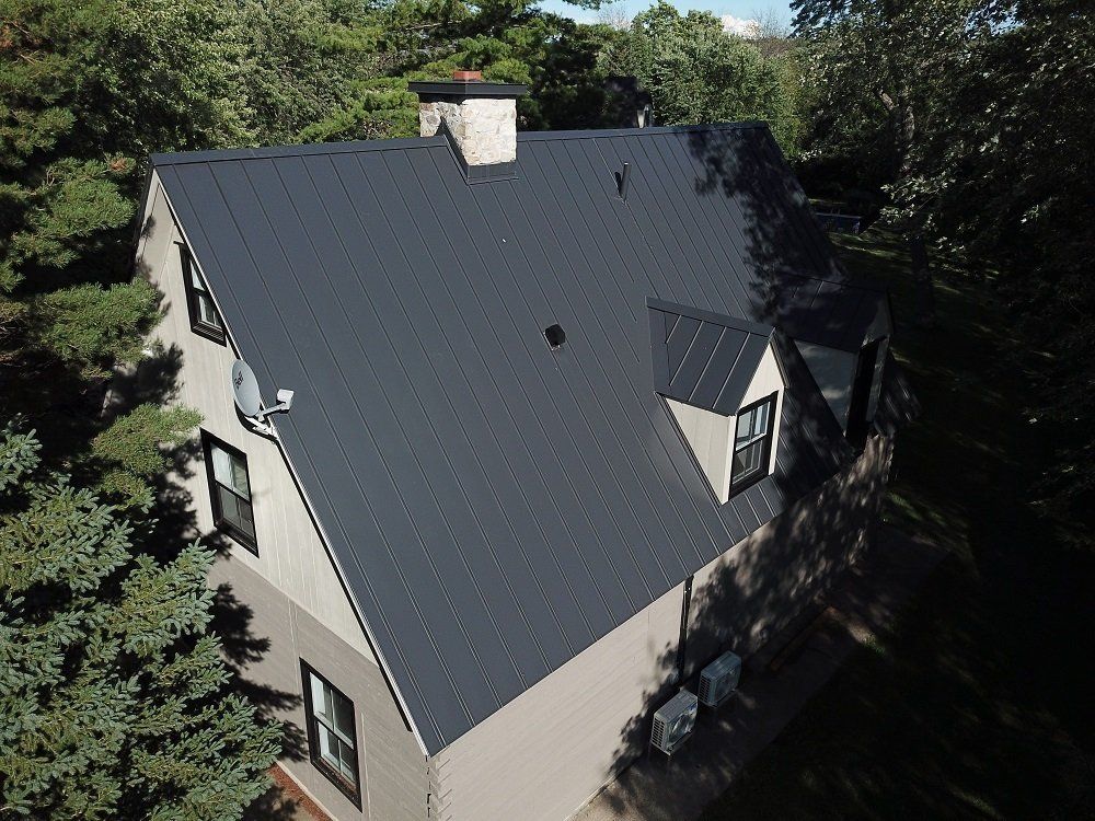 An aerial view of a house with a dark metal roof, beige siding, a stone chimney, and a dormer window among trees.