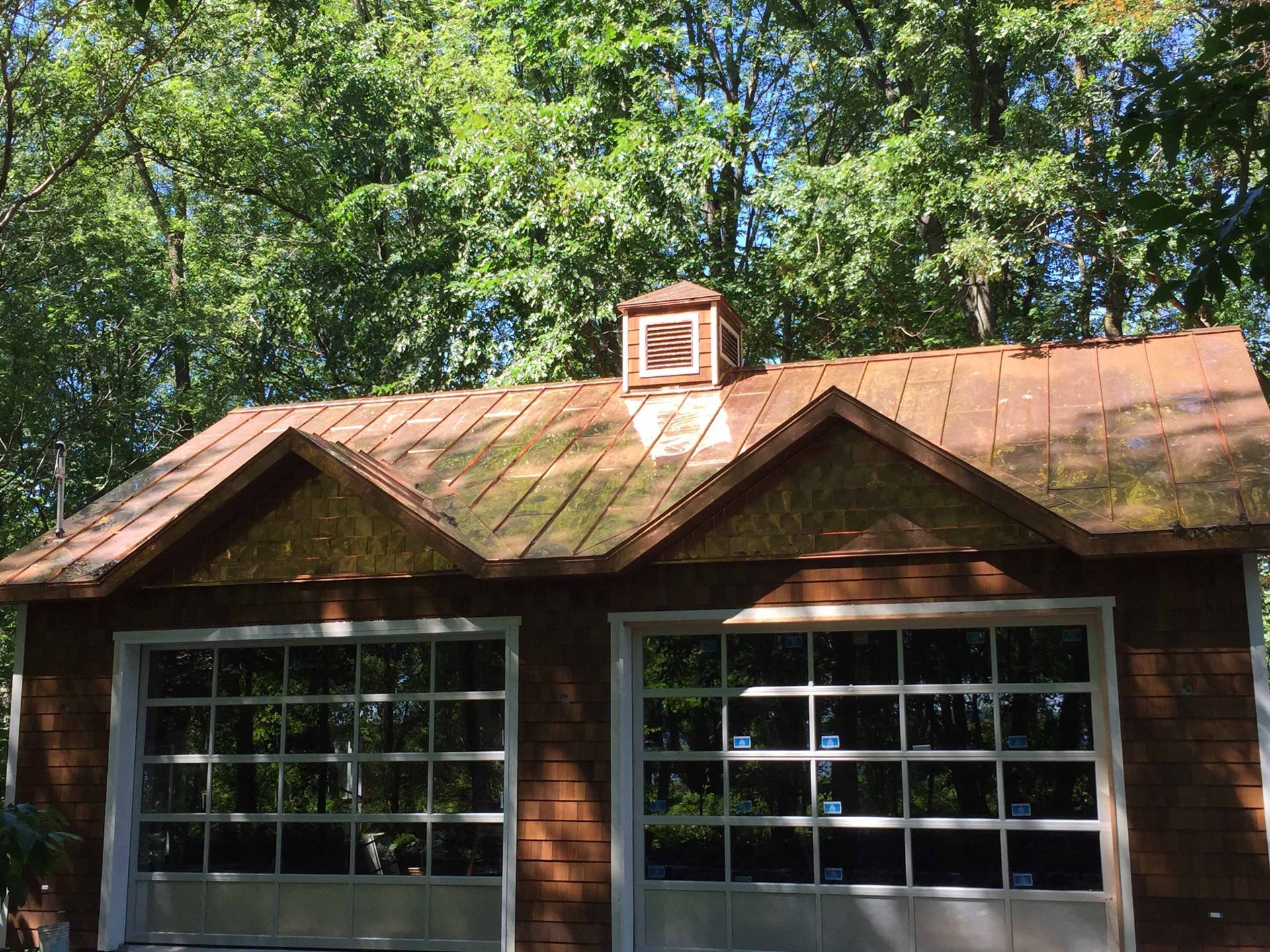 A detached wooden two-car garage with two glass garage doors, a double-gabled roof, and a small cupola amidst trees.