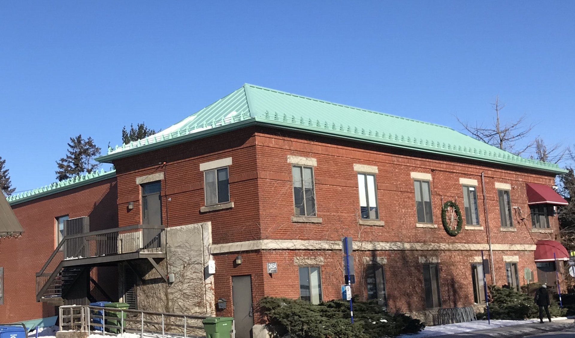 Two-story red brick building with a green roof, exterior wooden staircase, and seasonal wreath on a sunny winter day.