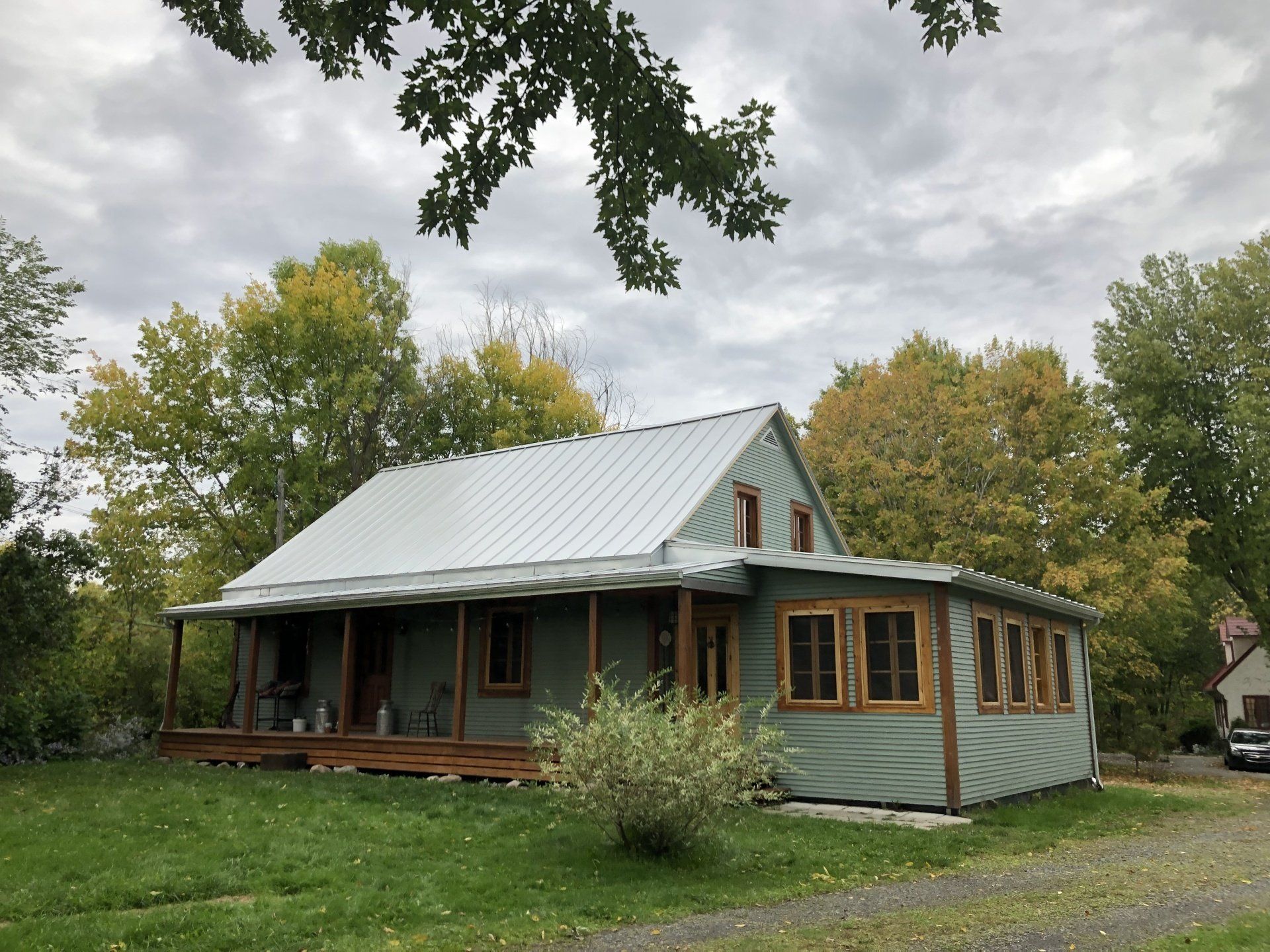 A pale green house with a silver metal roof, a wooden front porch, and autumn trees in the background.