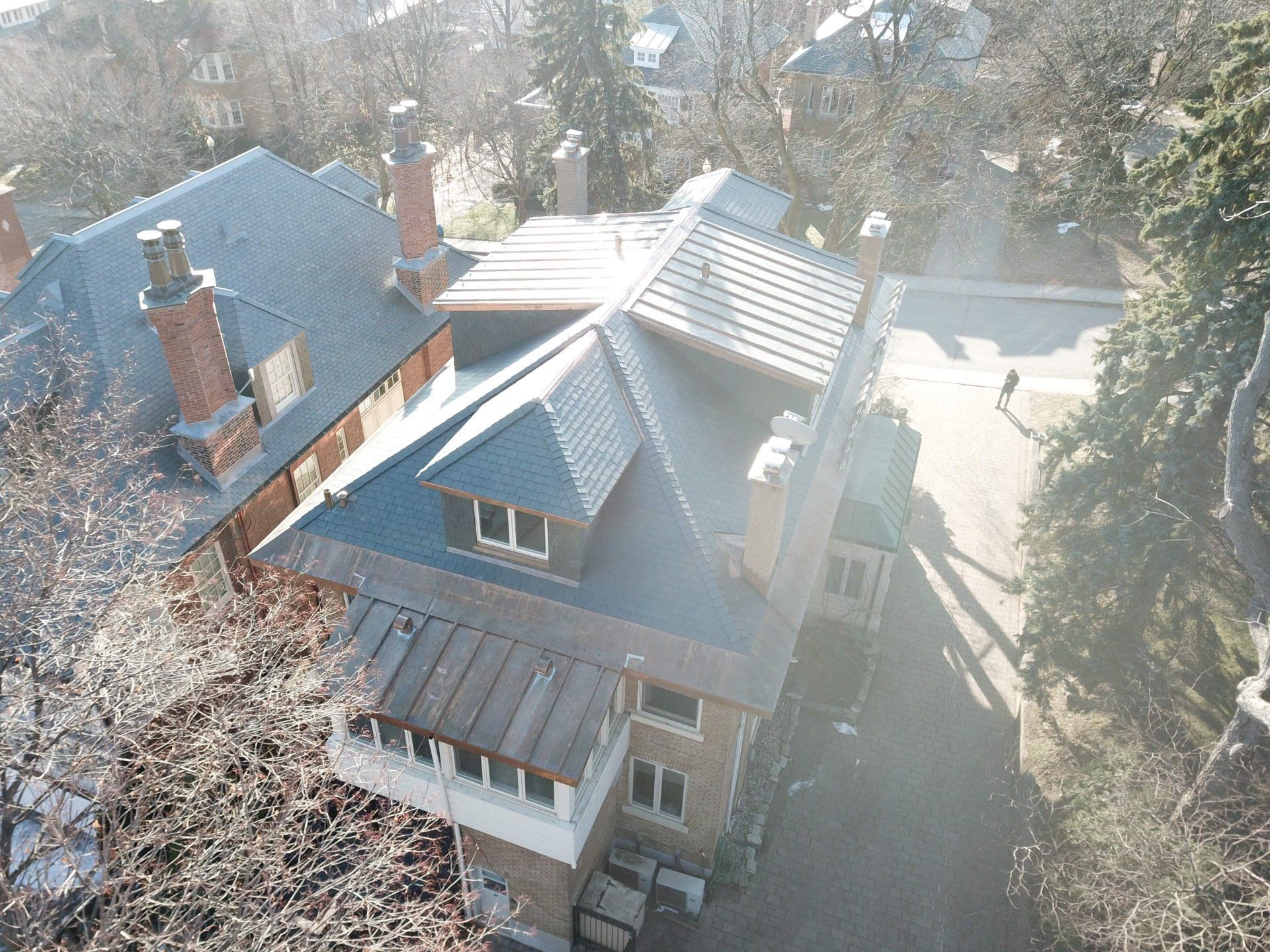An aerial view of a multi-story house with a grey slate roof, brick chimneys, and a dormer window on a sunny day.