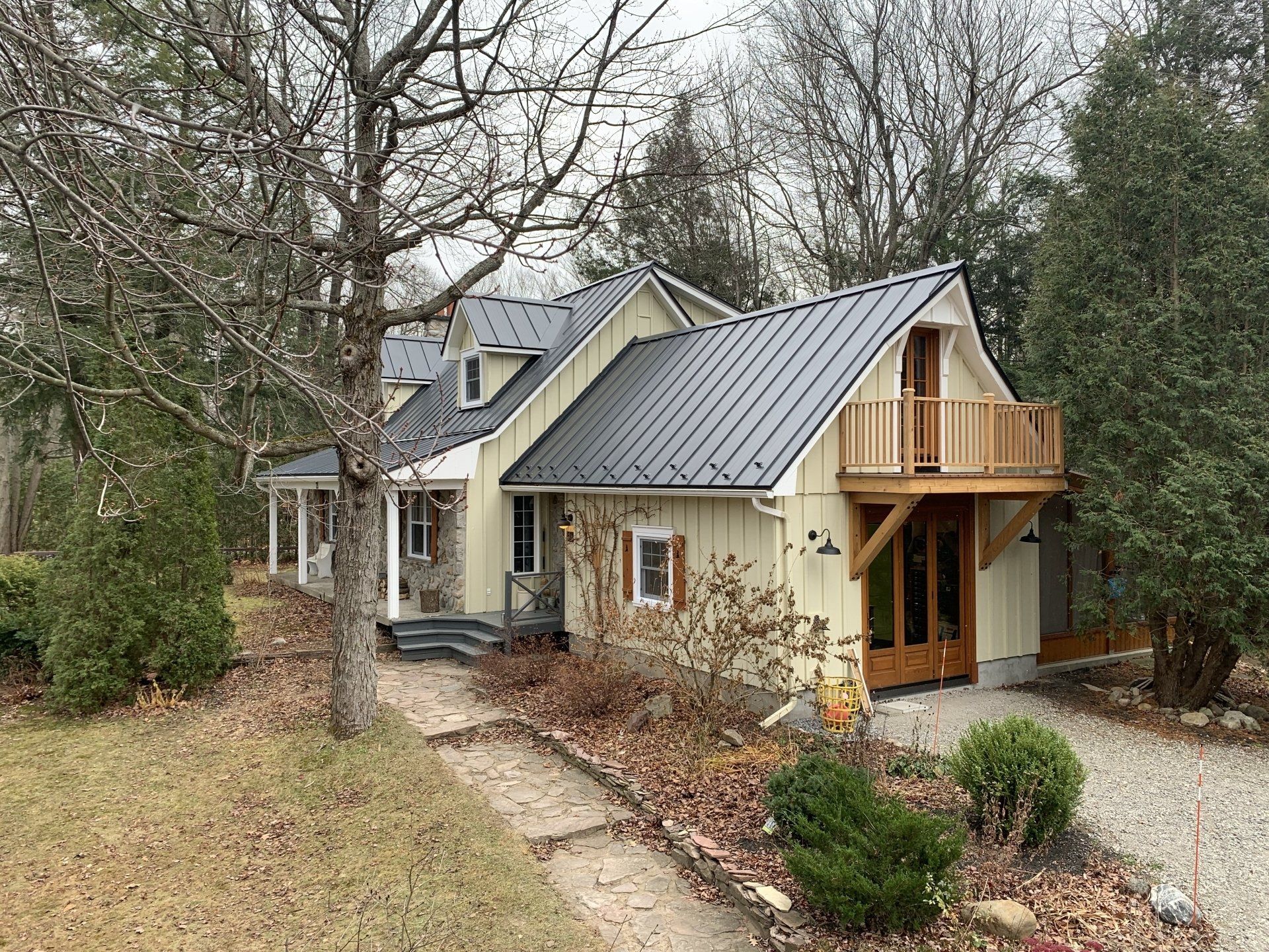 A light yellow two-story house with a black metal roof, front porch, and a small wooden balcony on a wooded lot.