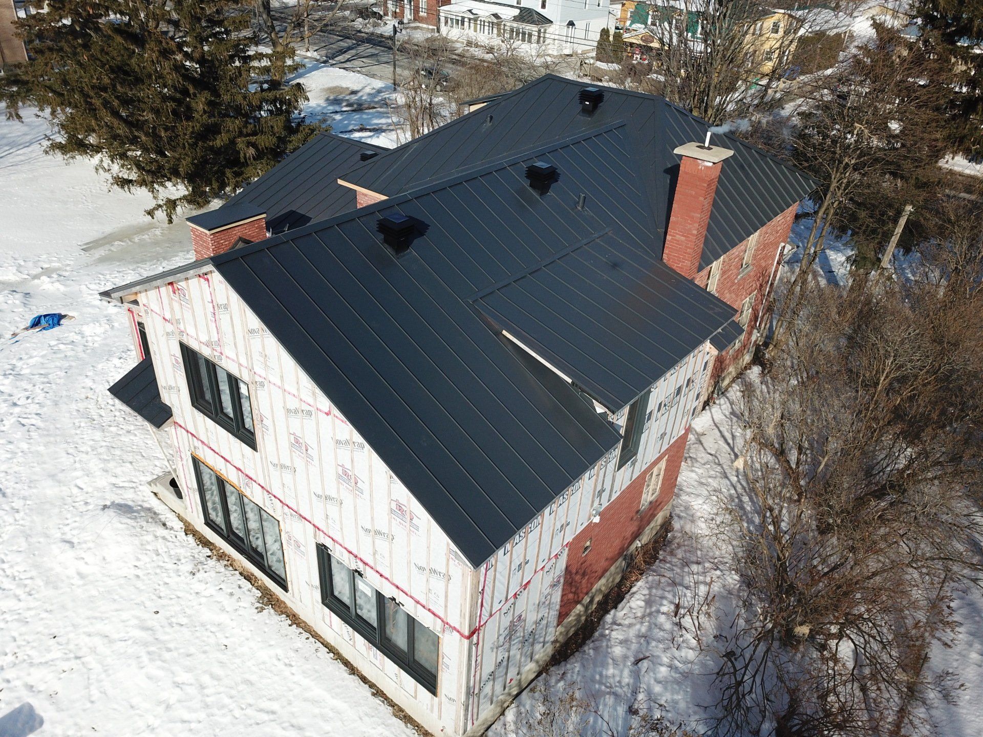 An aerial view of a brick house under construction, featuring a newly installed dark metal roof and snow-covered yard.