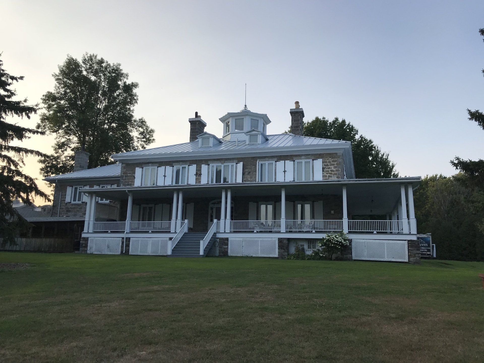A large, historic stone house with a wraparound porch, cupola, and metal roof set on a grassy lawn at sunset.