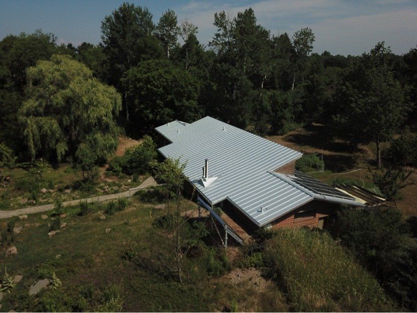 Aerial view of a house with a gray metal roof, chimney, and solar panels nestled in a lush, wooded landscape.