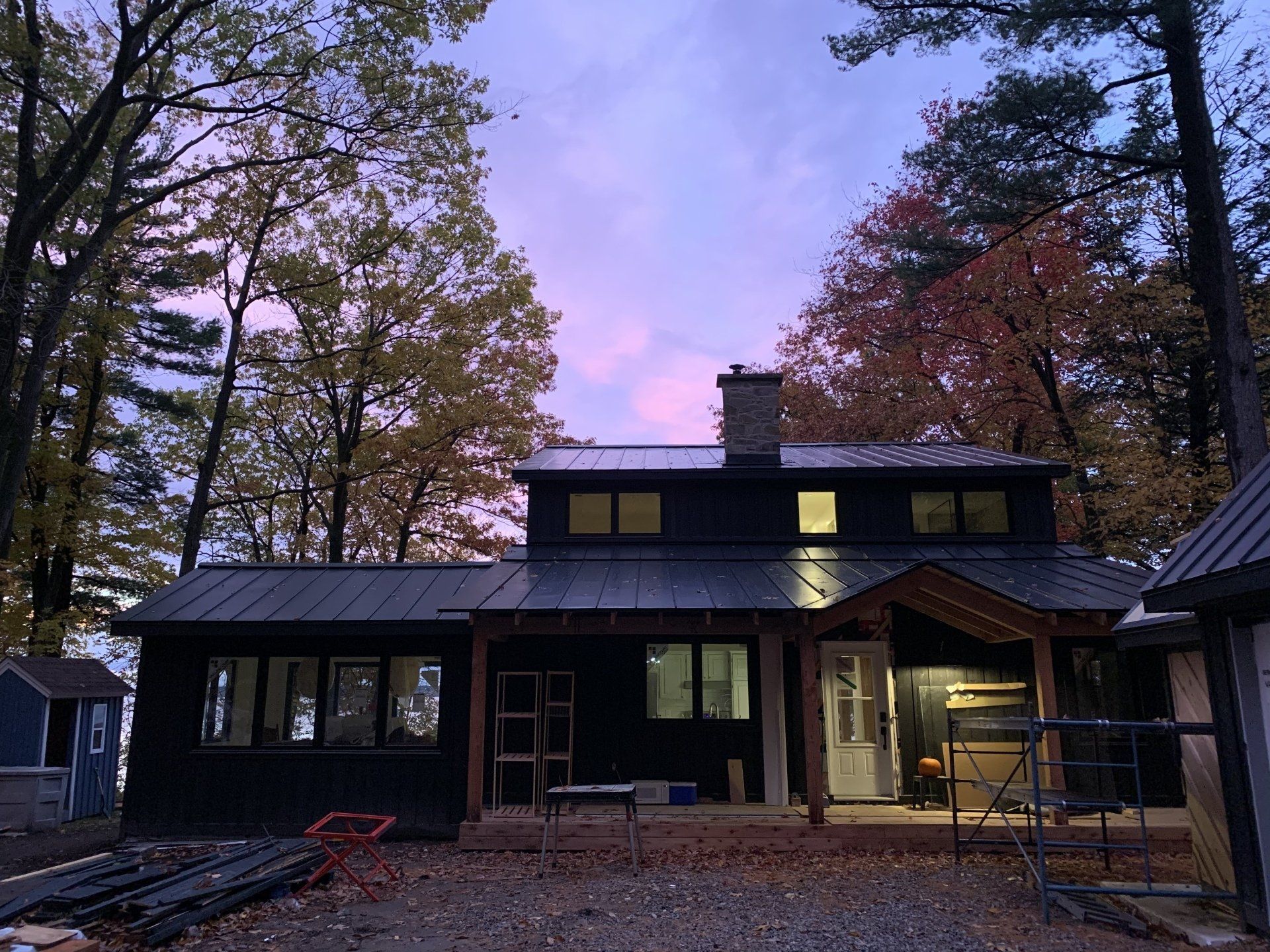 A dark-sided two-story house with a metal roof and a wooden porch at dusk, surrounded by autumn trees under a pink sky.