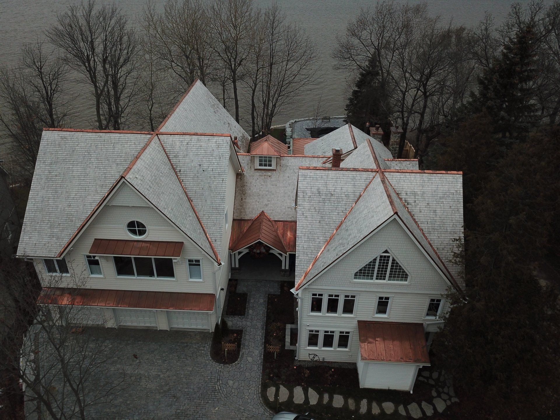 An aerial view of a large white house with copper-accented roofs, situated by a body of water in a wooded setting.