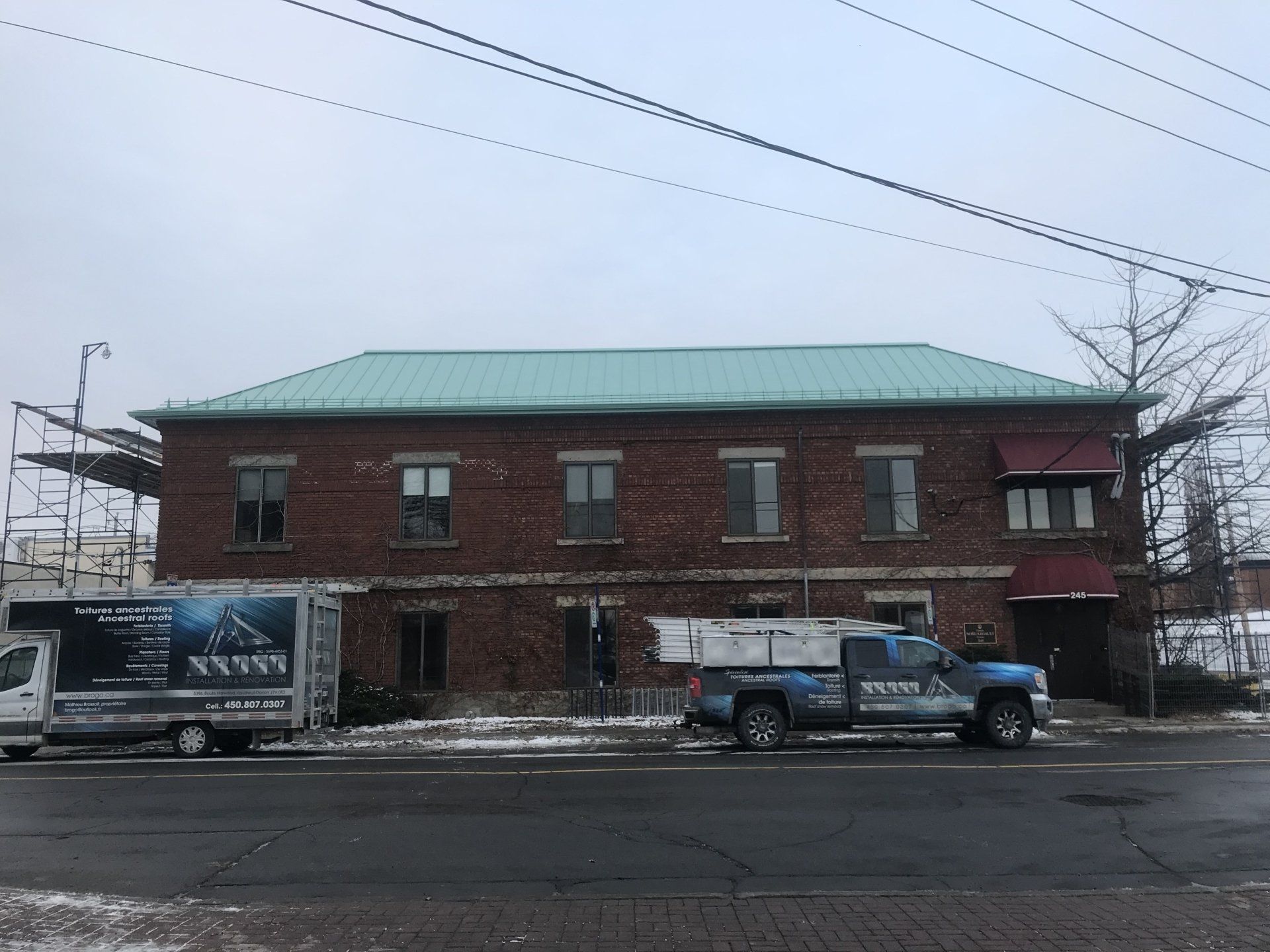 A brick two-story building with a light green roof, with work trucks parked in front on a street on an overcast day.