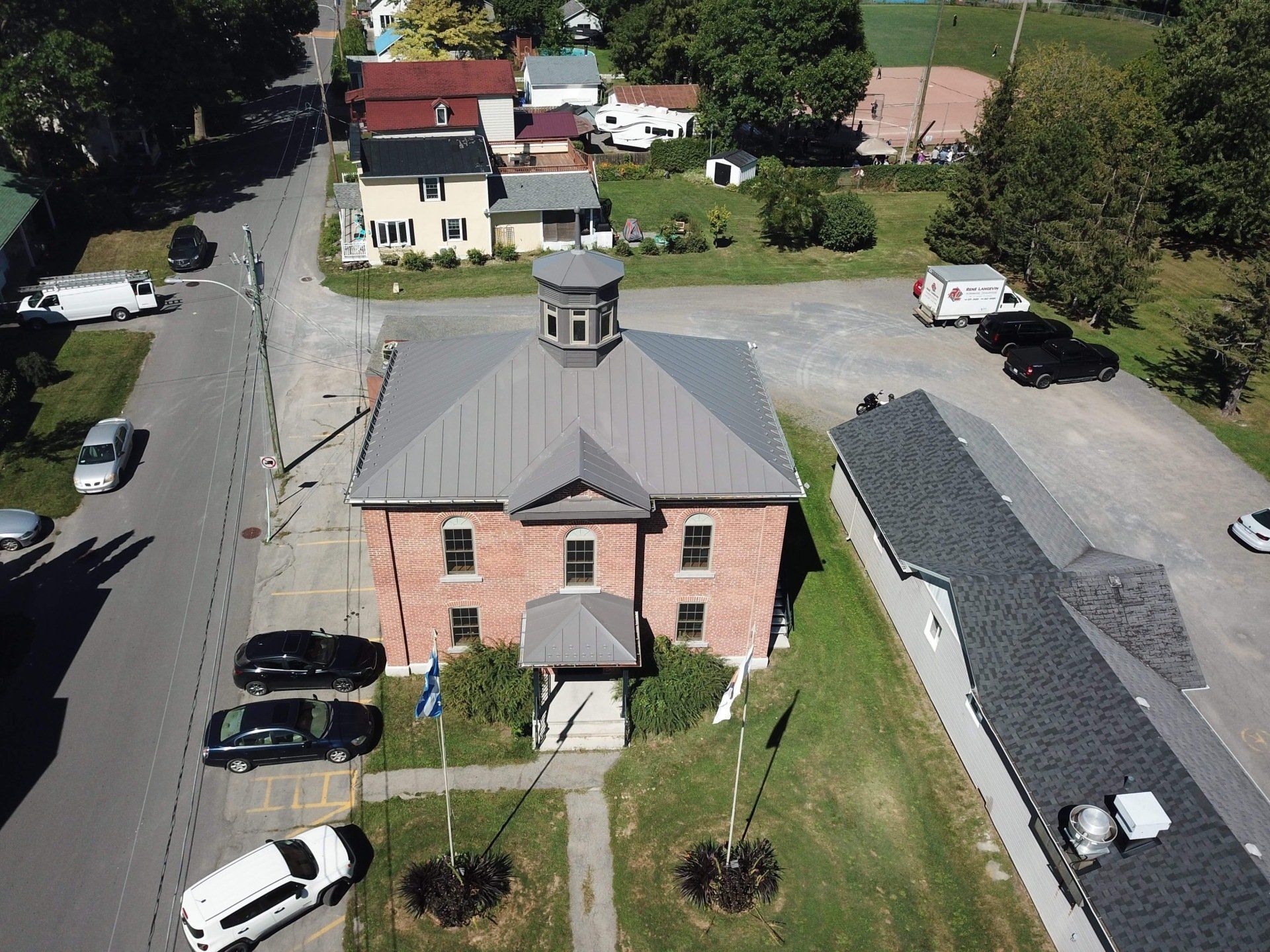 Aerial view of a red brick municipal building with a gray metal roof, cupola, and adjacent parking lot.