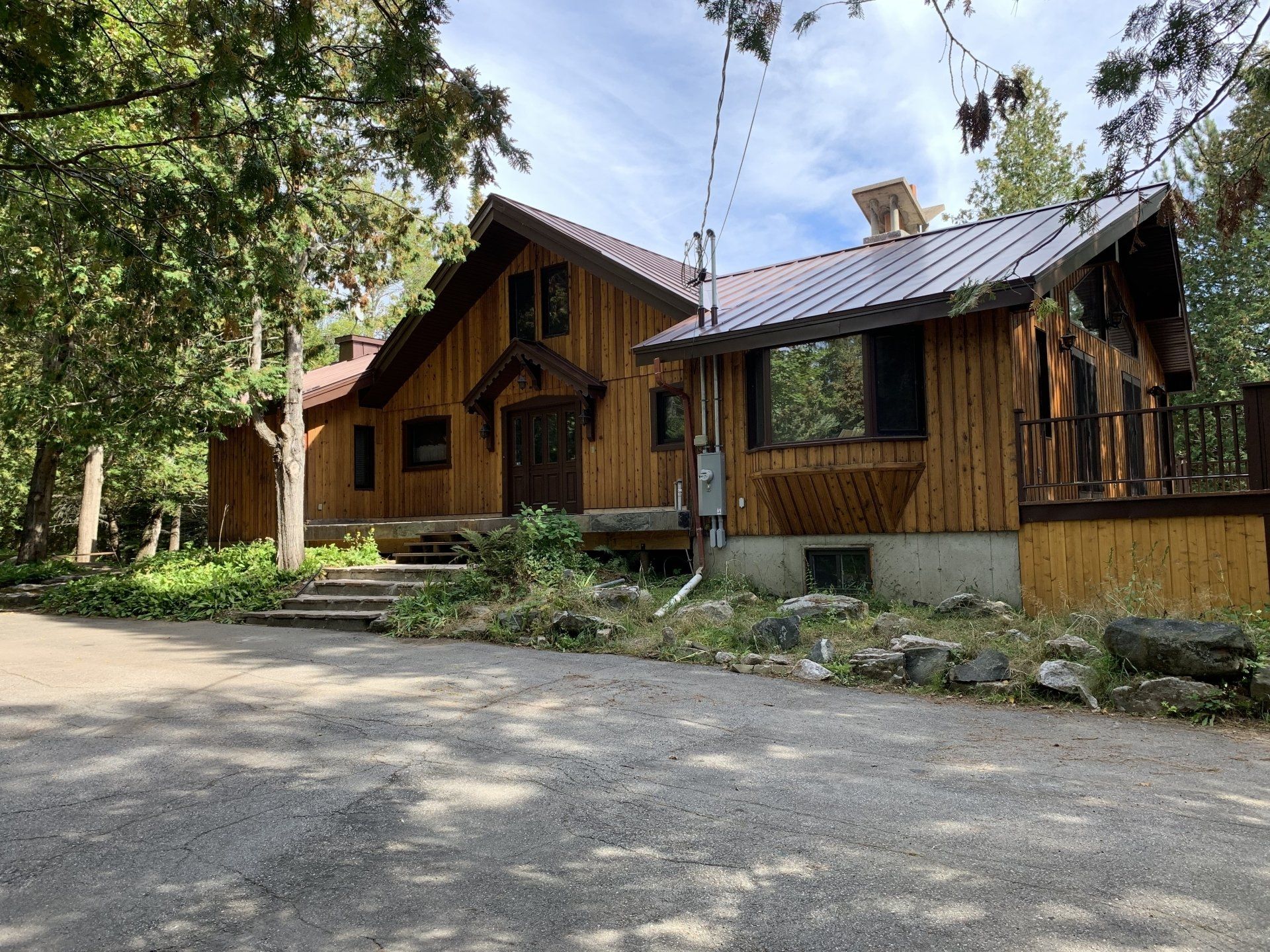 A two-story log-style cabin with a metal roof surrounded by trees and a gravel driveway in a natural, wooded setting.