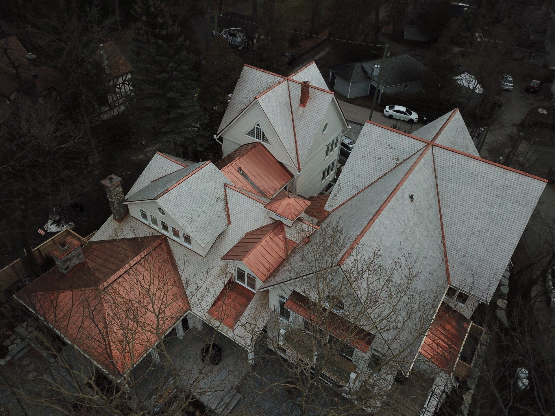 High-angle aerial view of a large residential house with a complex roof made of light grey shingles and bright copper.