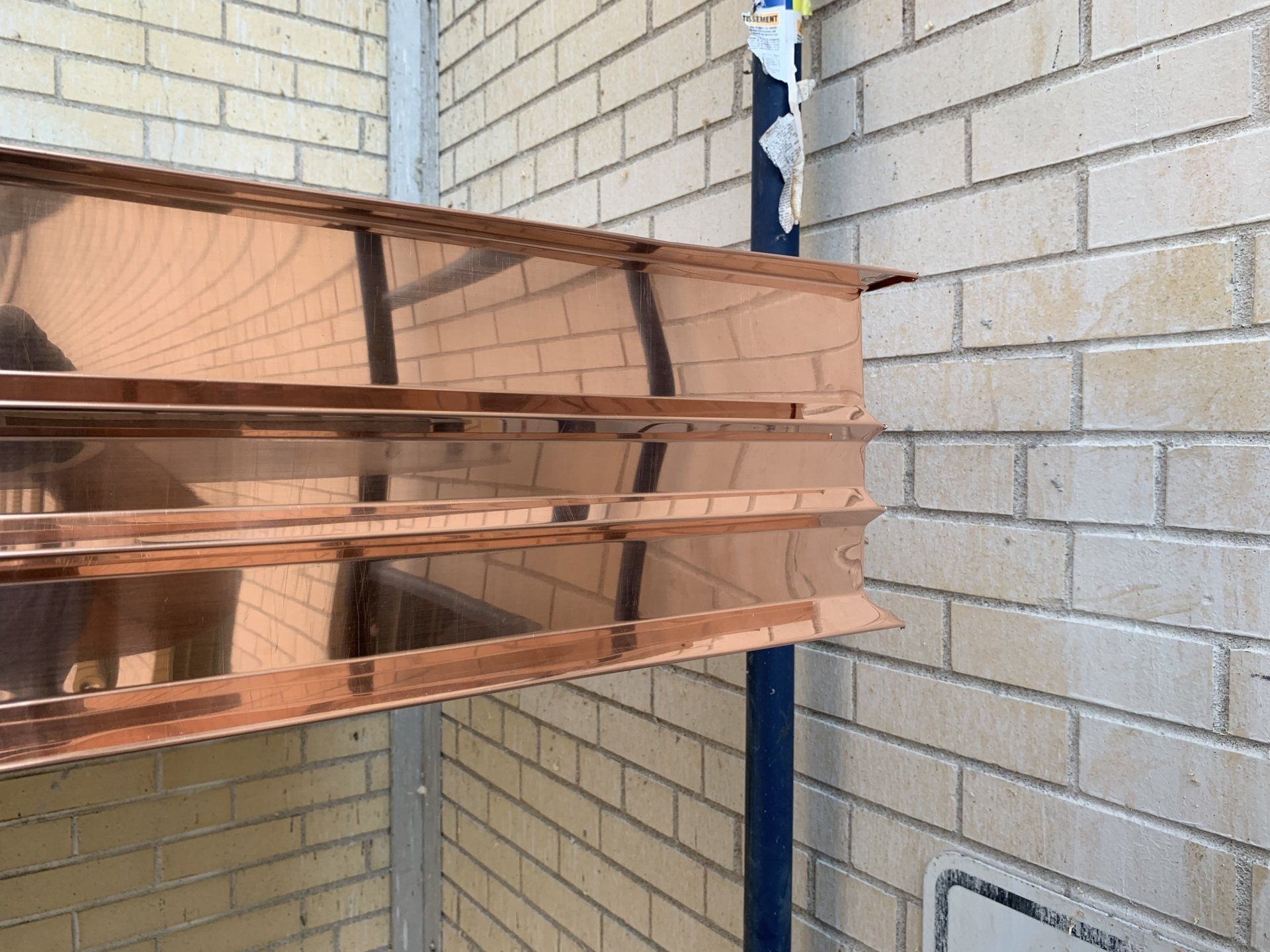 A close-up of a polished copper-colored architectural panel with horizontal grooves, mounted against a light brick wall.