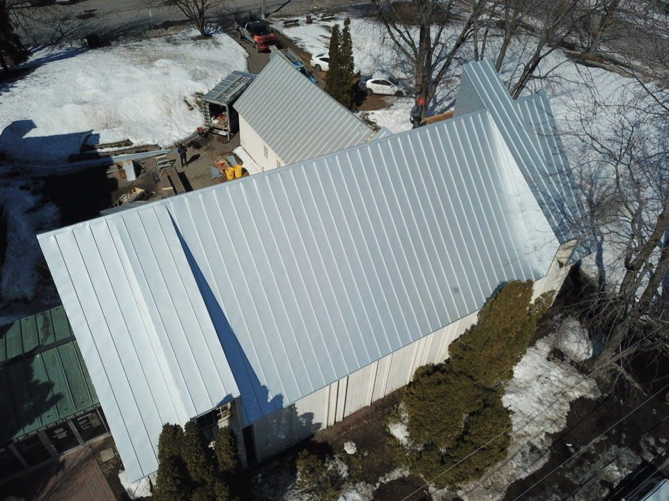 An aerial view of a building with a light-colored metal roof, surrounded by trees and patches of melting snow.