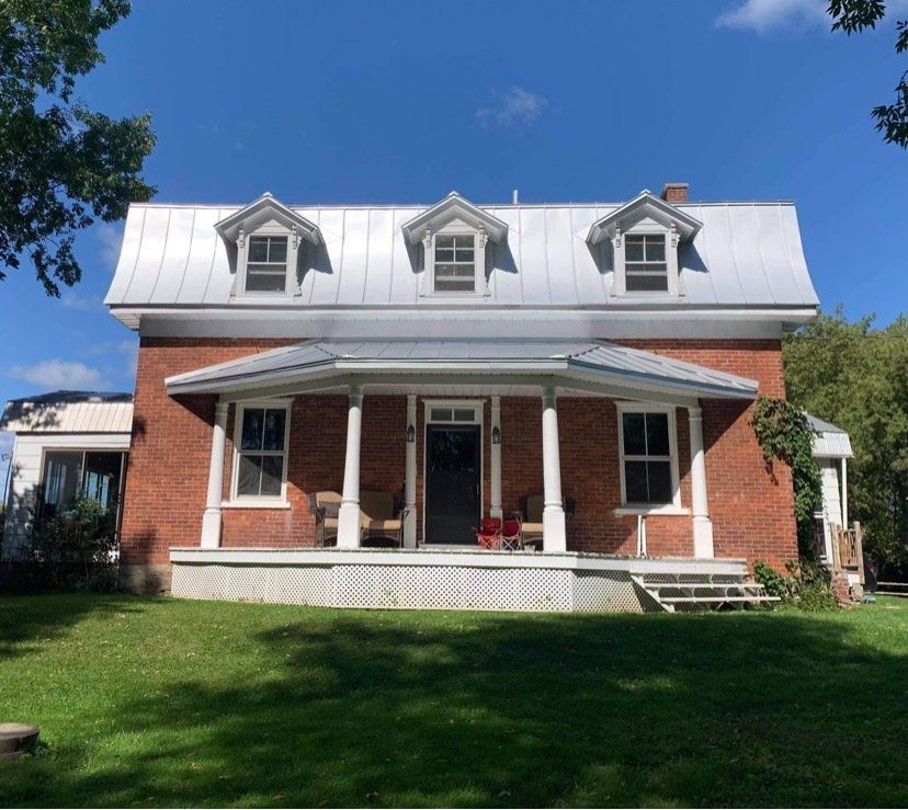 A two-story brick house with a metal gambrel roof, dormer windows, and a covered front porch, set on a green lawn.