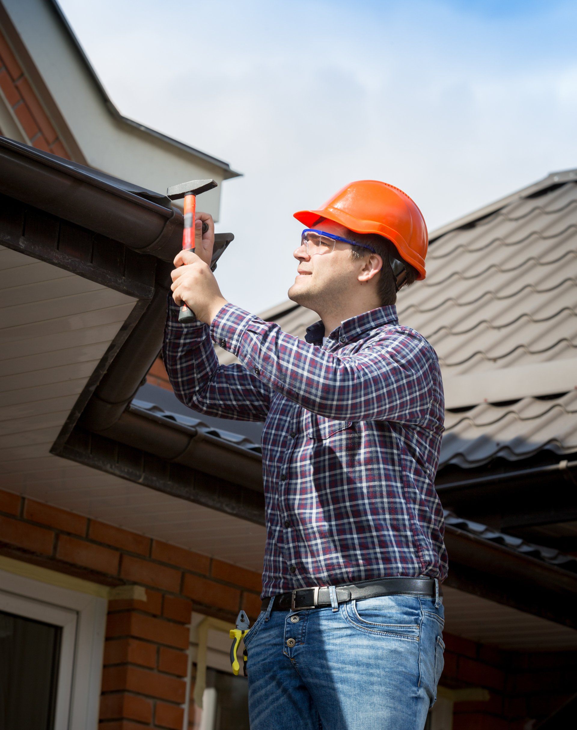 Une personne portant une chemise à carreaux et un casque de chantier orange utilise un marteau pour travailler sur la toiture d'une maison en briques.