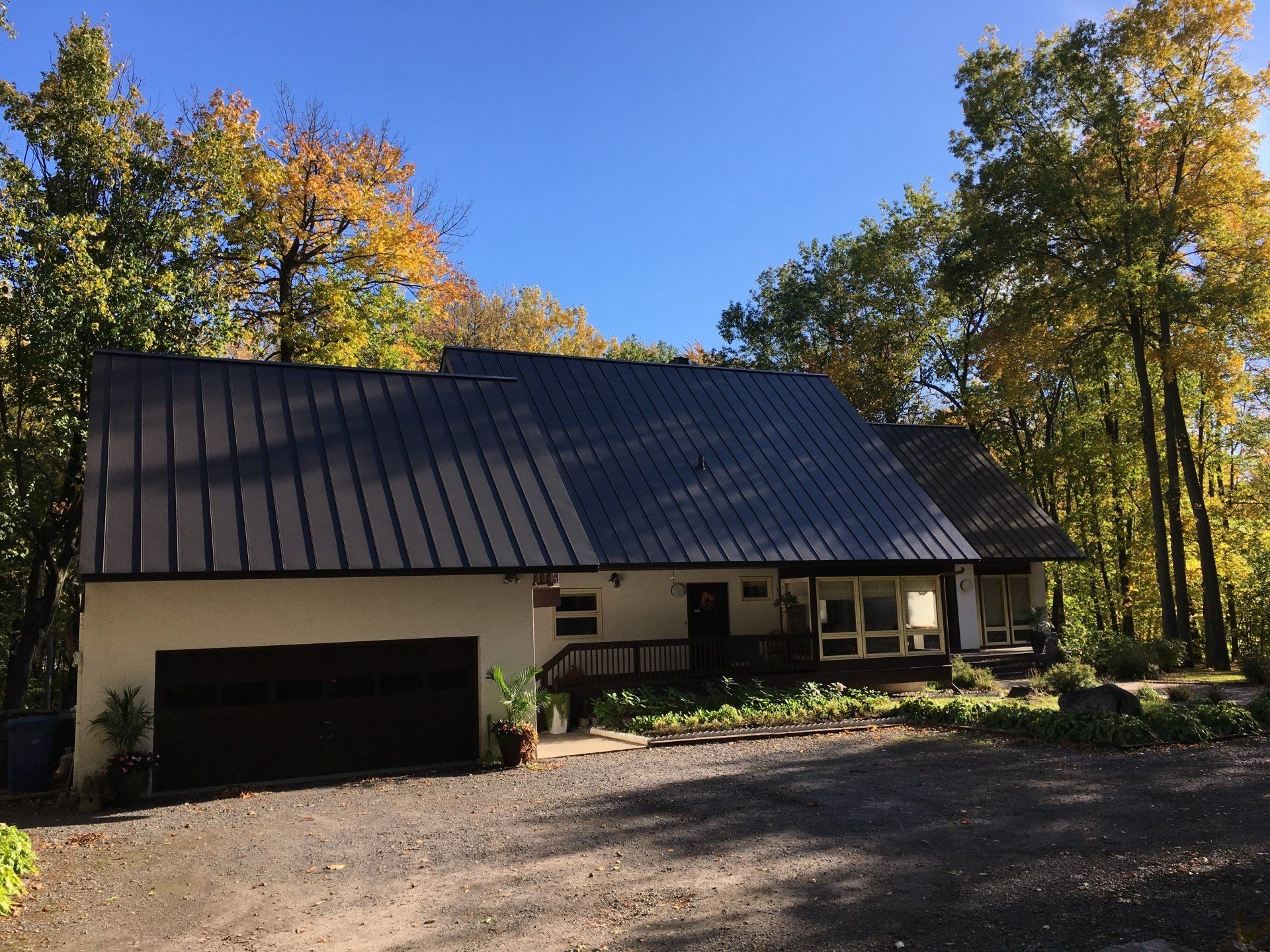 A white house with a dark metal roof, garage, and front porch surrounded by trees with autumn leaves.