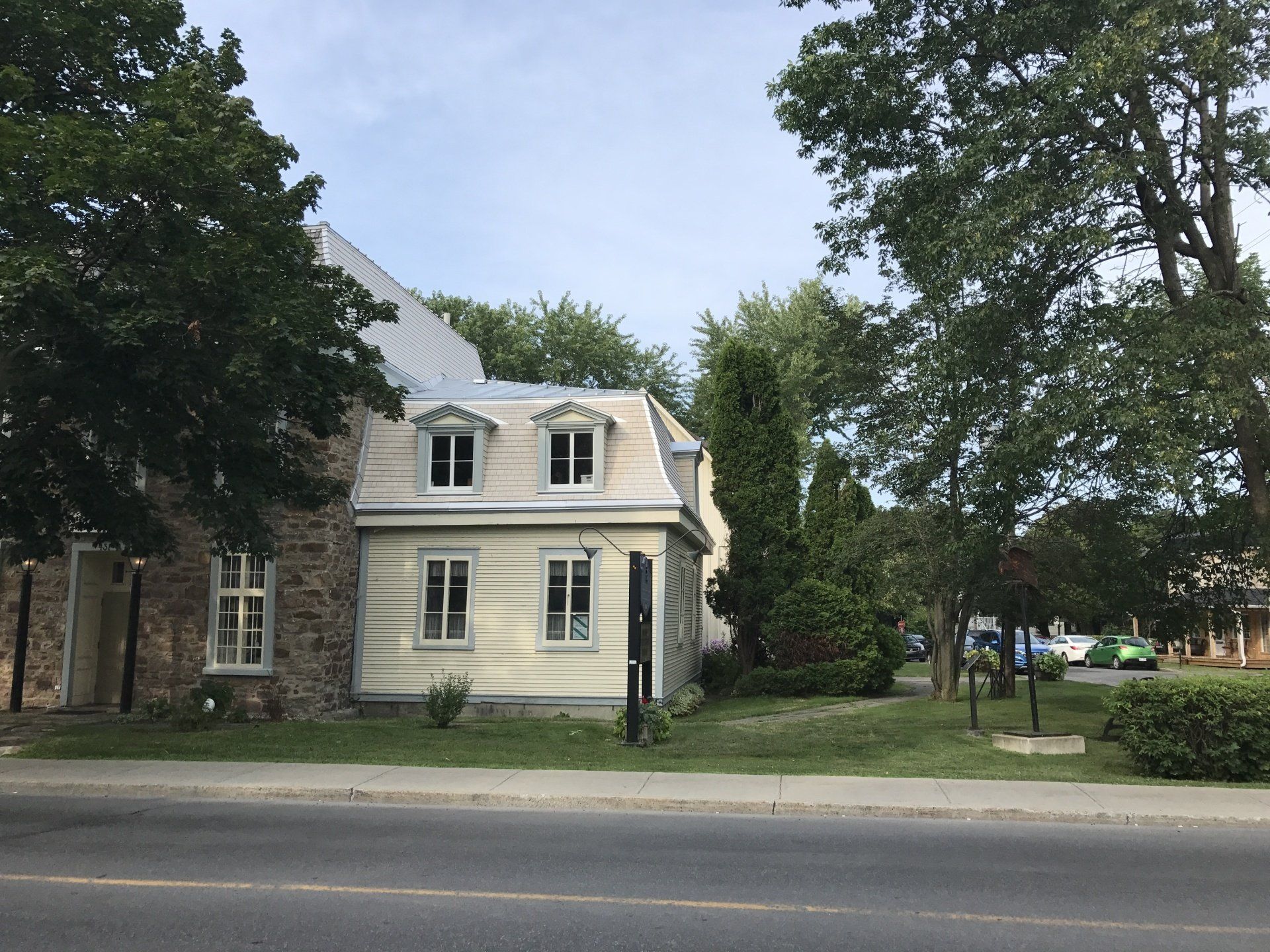 A two-story building with stone and light-colored wood siding, dormer windows, and a metal roof, framed by mature trees.