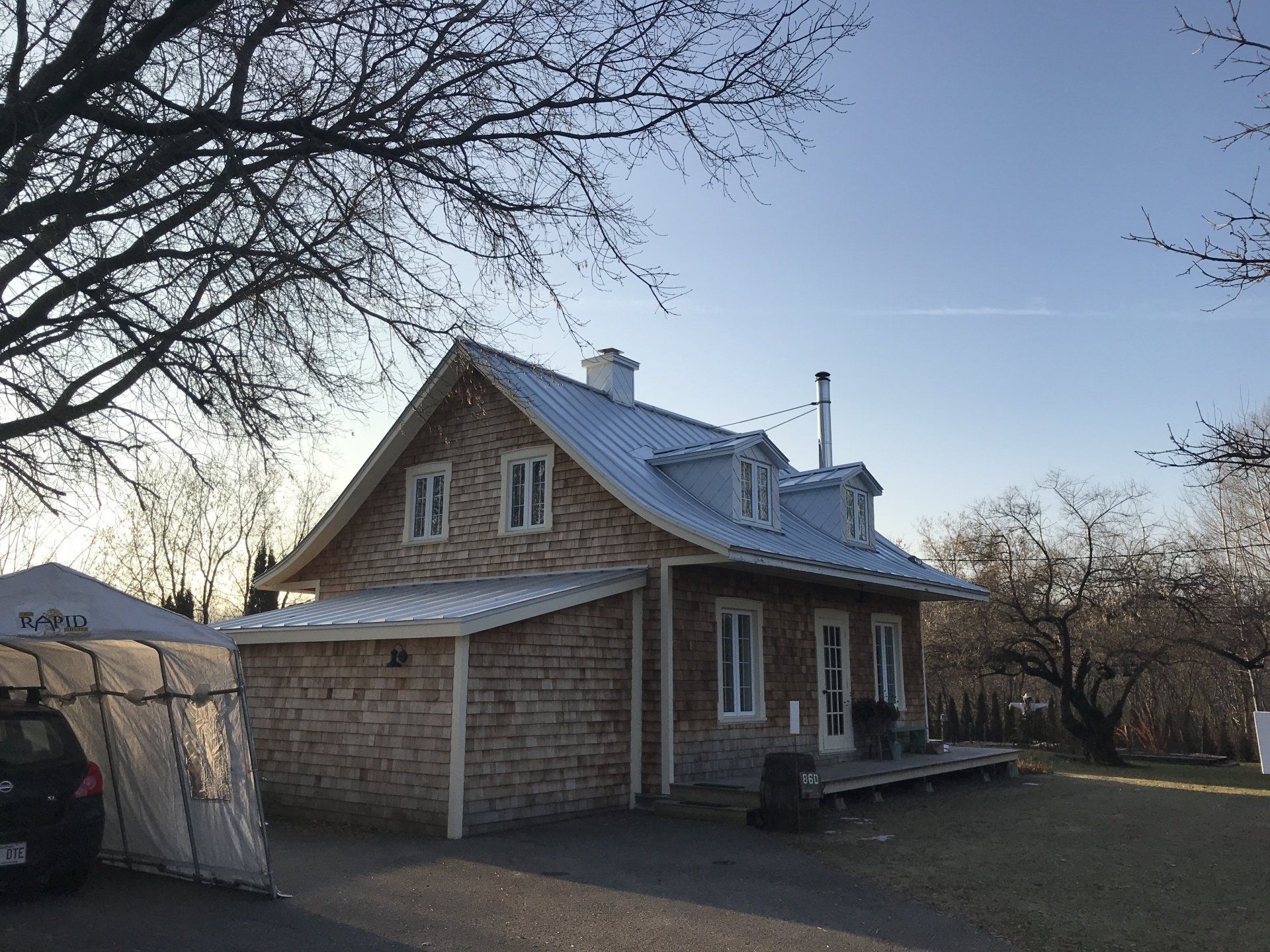 A stone house with a metal roof and dormer windows, situated in a yard with trees and a portable car shelter.