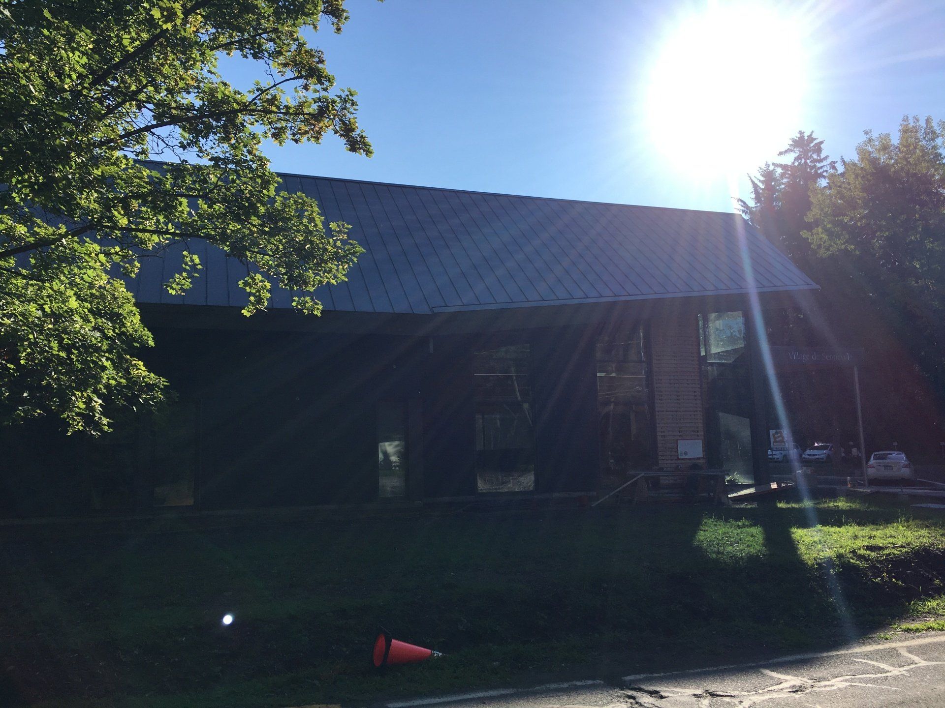 A side view of a building with a gray metal roof, partially shaded by large trees on a sunny day.
