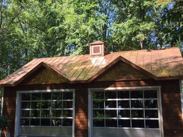 A rustic two-car garage with glass-paneled doors, a metal roof, and a small vent cupola, surrounded by lush trees.