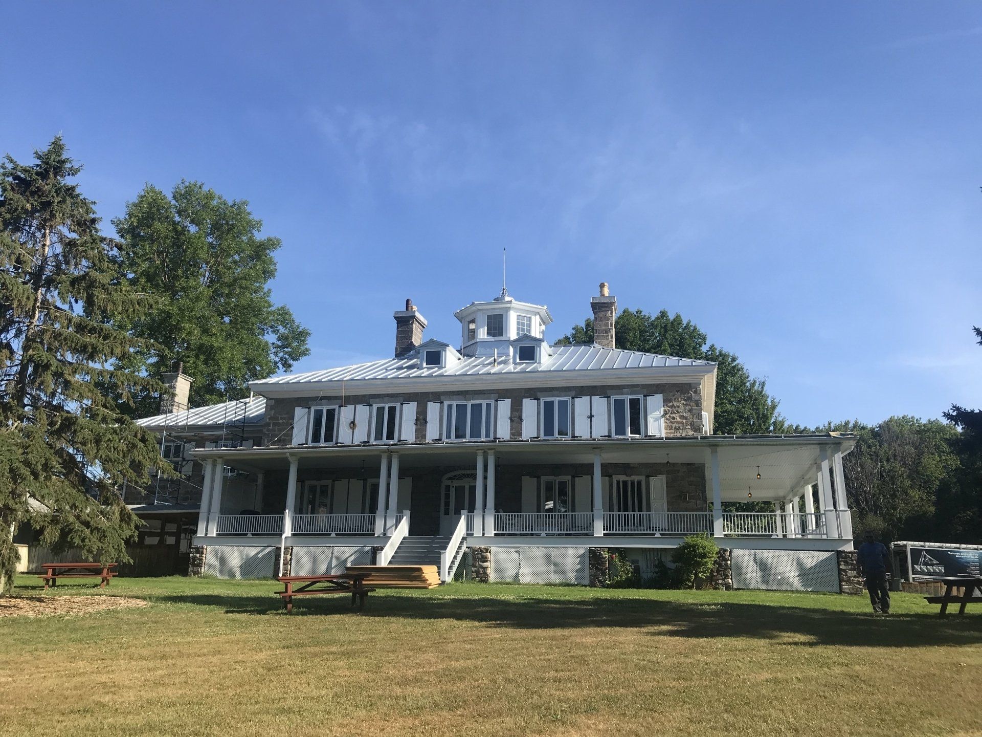 A historic two-story stone house with a wide front porch, white shutters, and a cupola under a clear blue sky.