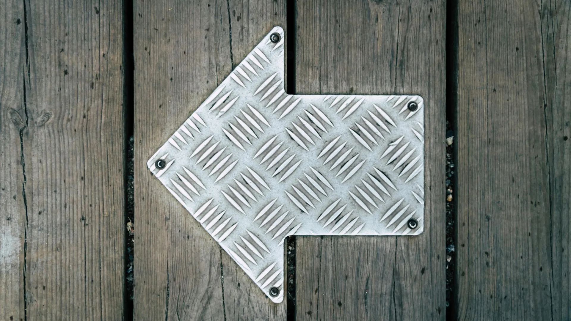 Silver metal arrow sign pointing left on weathered wooden planks.