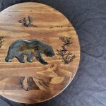 Wooden round with carved bear and desert landscape.