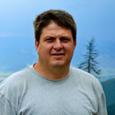 Portrait of a man in a light gray T-shirt standing outdoors with blue sky and mountains behind him