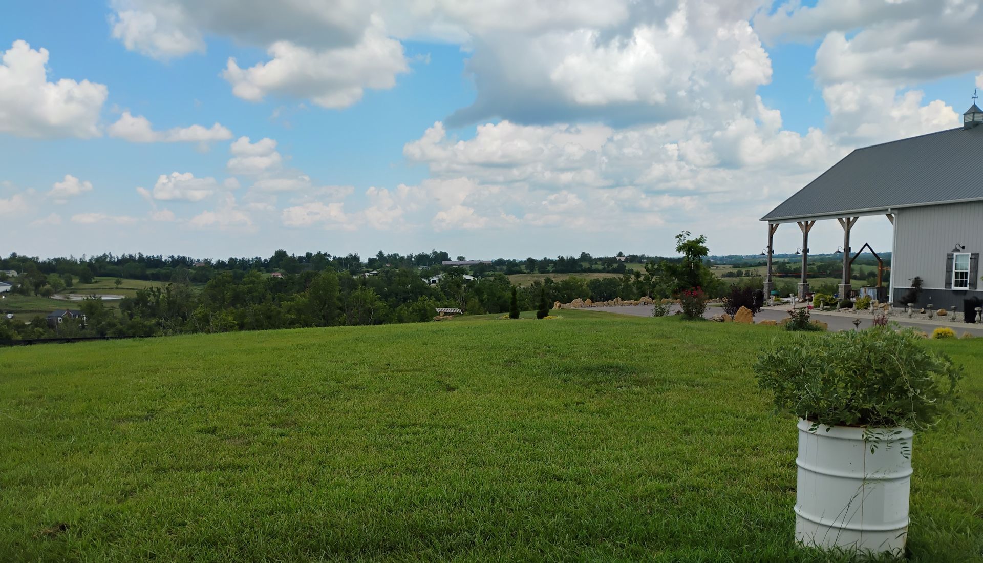 Wide grassy field under a blue sky with clouds, next to a house and a potted plant on the right.