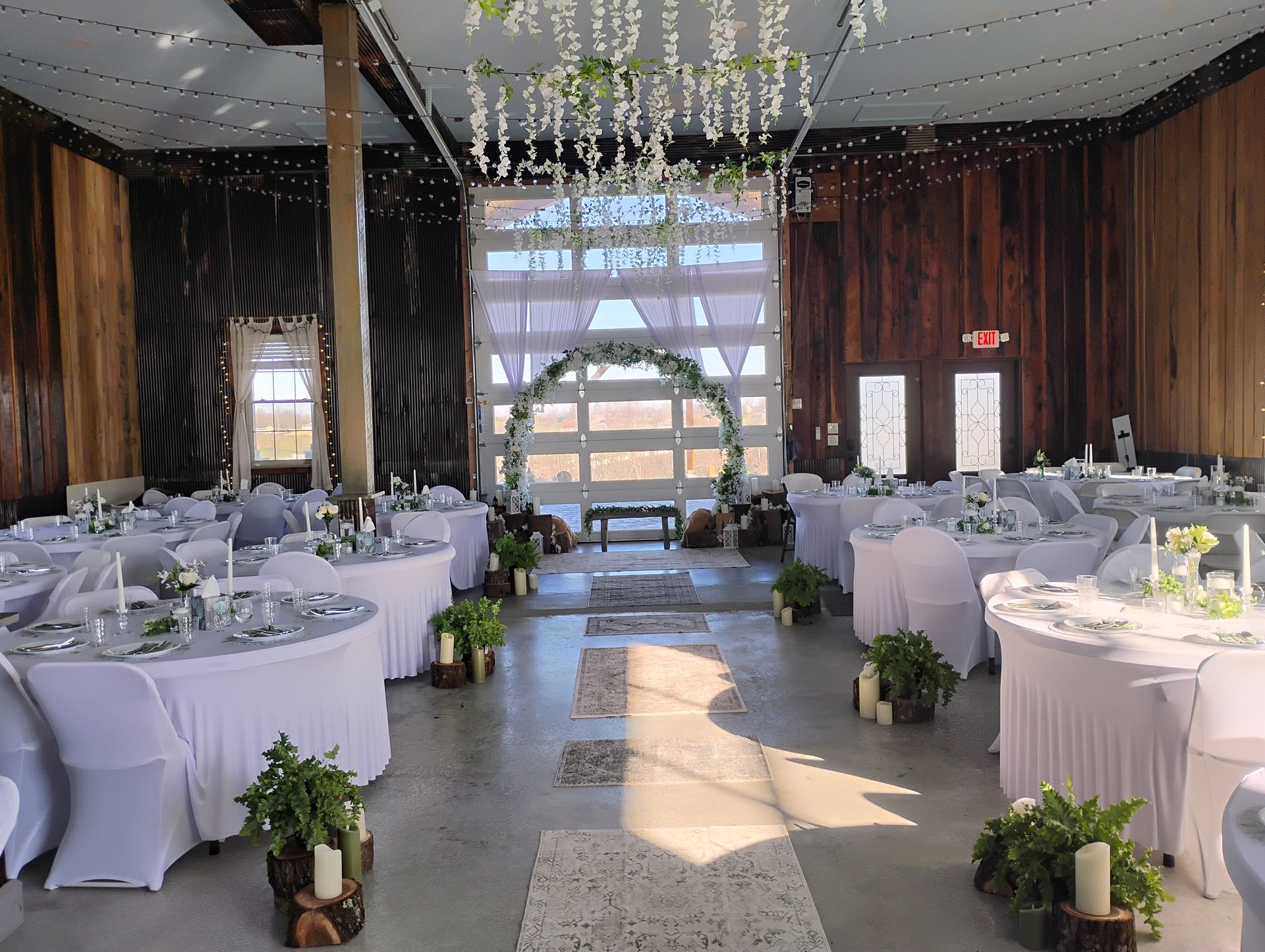 Elegant banquet hall with round white-clothed tables, greenery, and sunlight through a large arched window