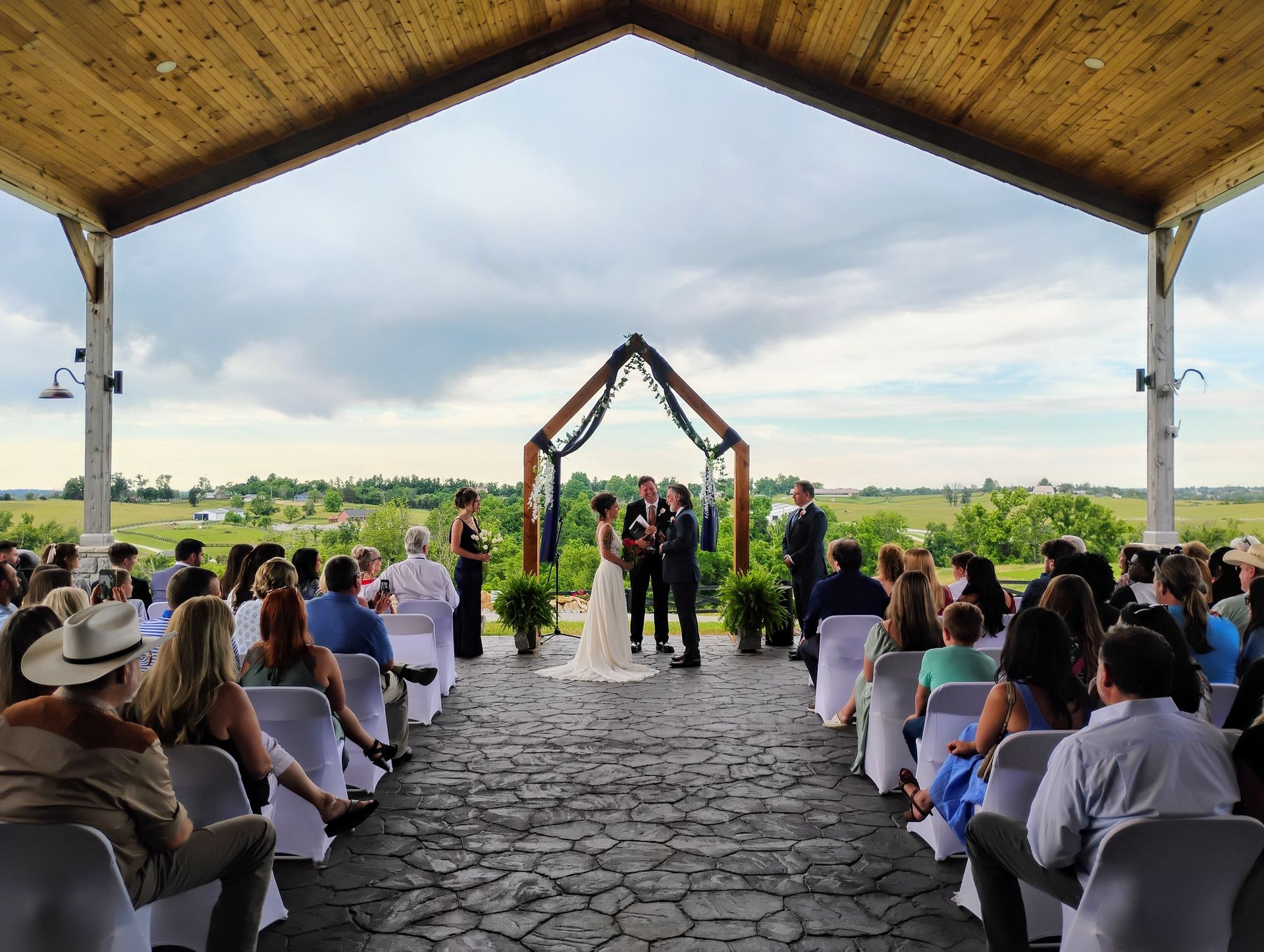 Wedding ceremony under a wooden pavilion with guests seated, a couple at the altar, and a green field beyond.