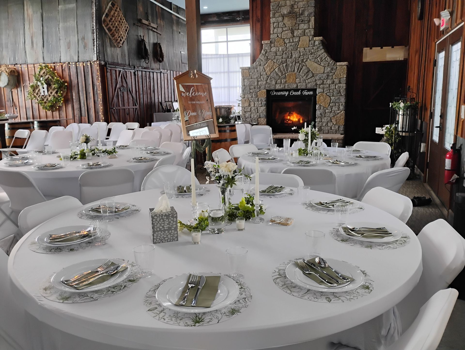 Round banquet tables set for a wedding reception in a rustic hall, with a fireplace and white chair covers.