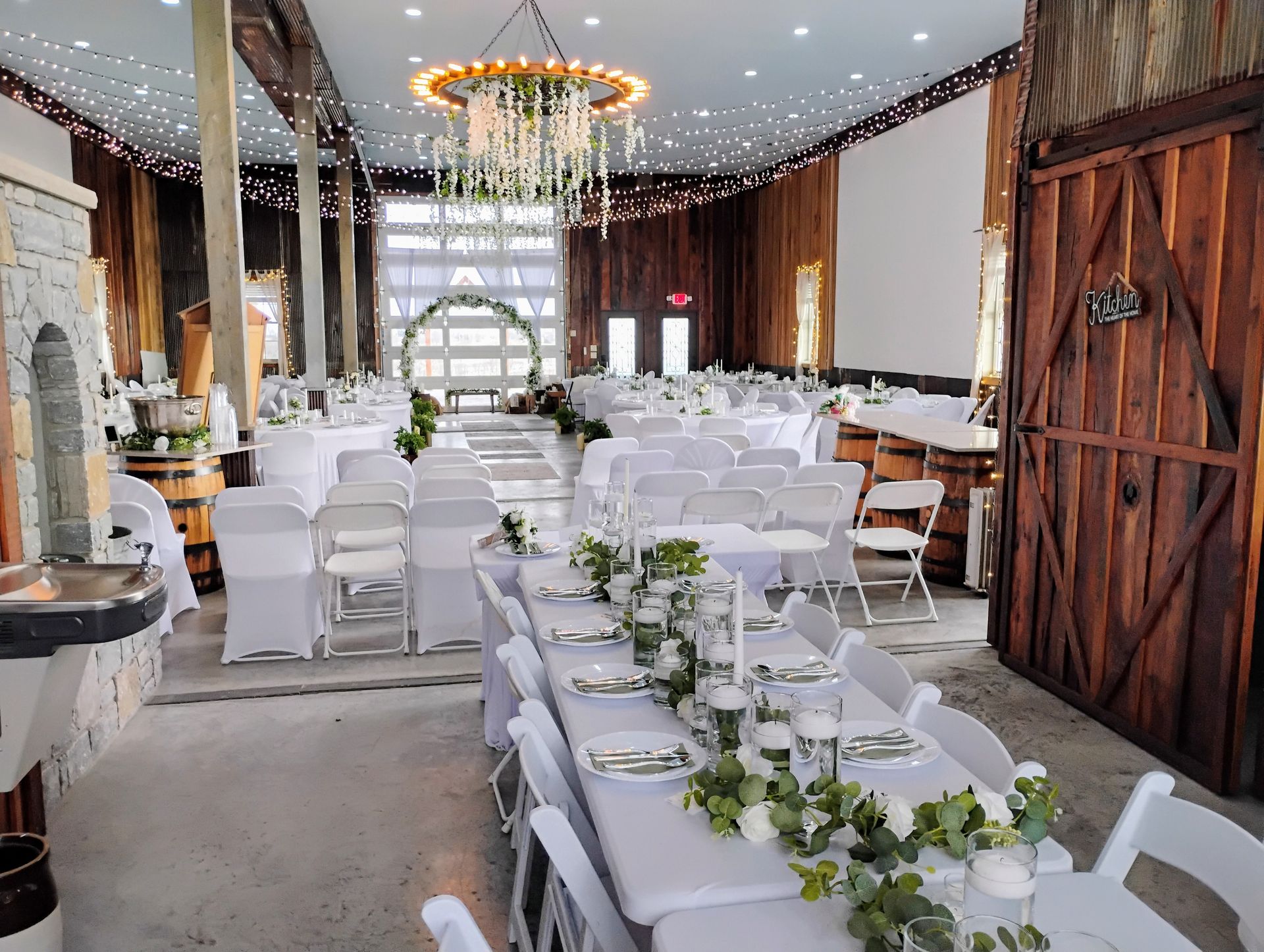 Decorated rustic banquet hall with white chairs and long tables set for a wedding reception