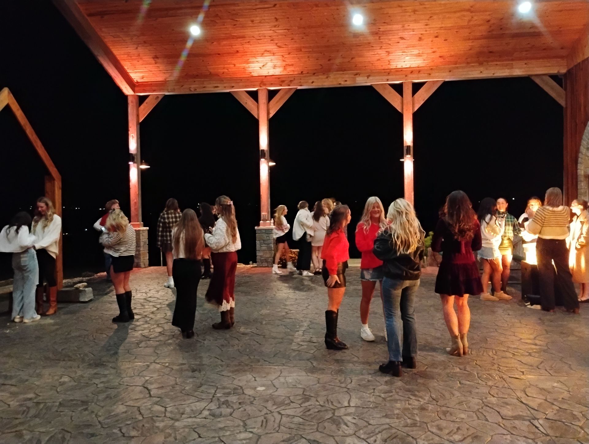 People dancing in a wooden pavilion at night, with warm lights and a stone floor.